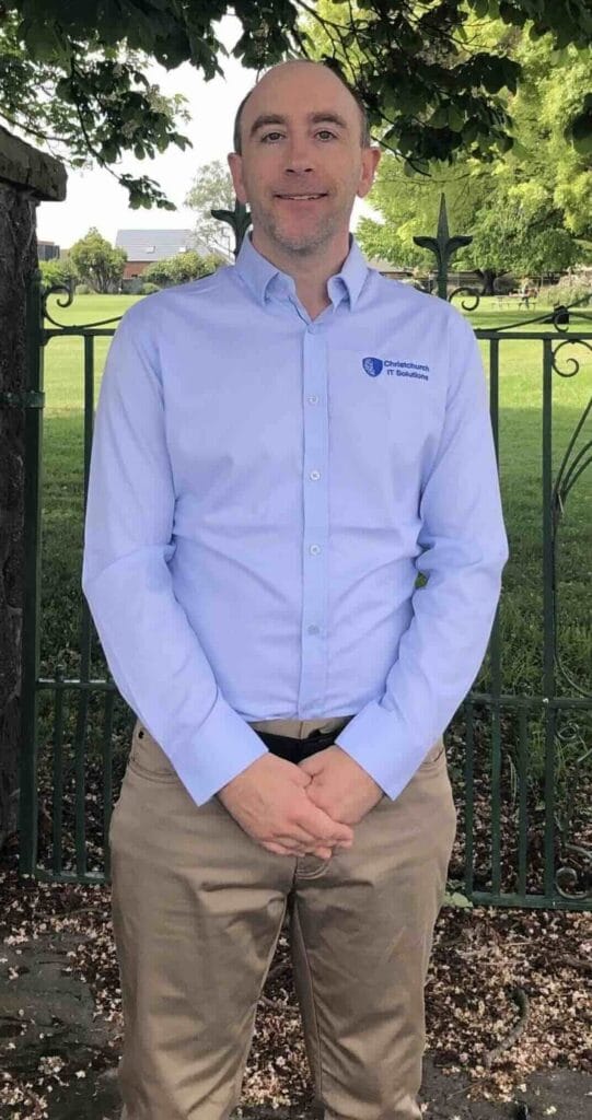 A person in a light blue shirt standing outside a stone gate with a green lawn and trees in the background.